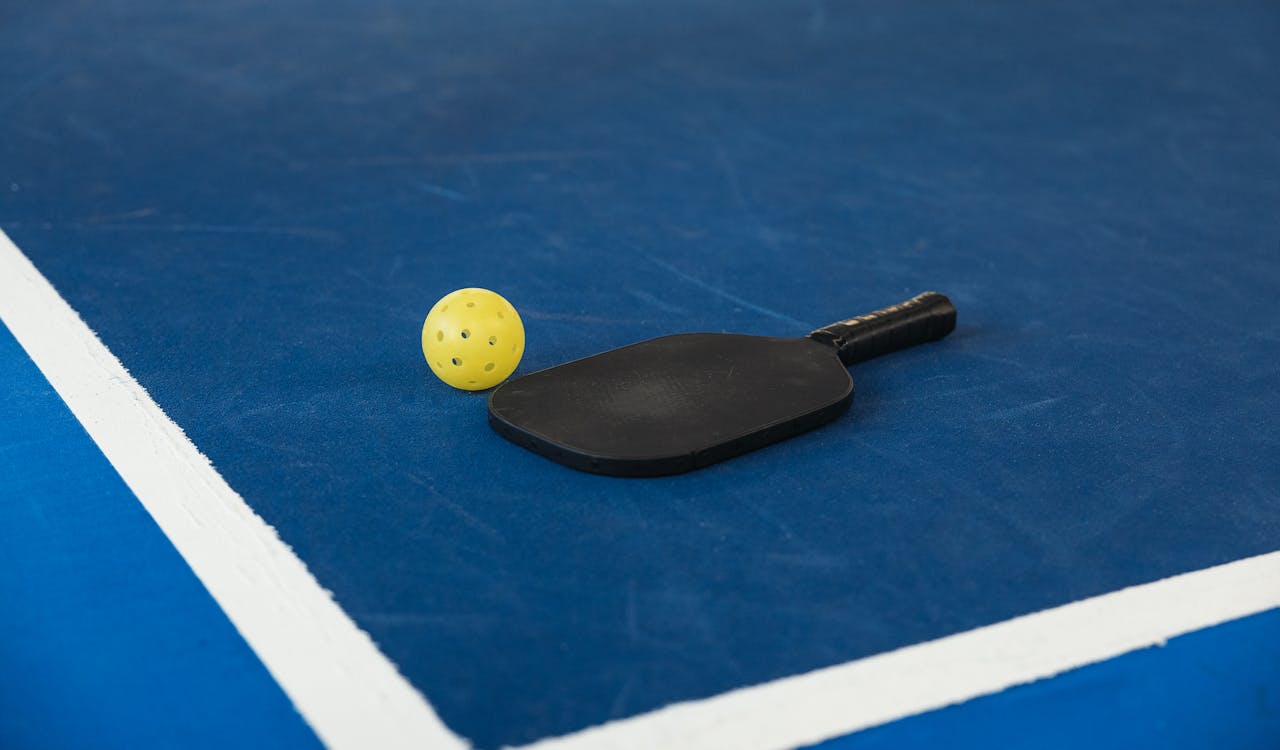 our-journey-01 A pickleball paddle and yellow ball on a blue court corner, symbolizing sport and activity.