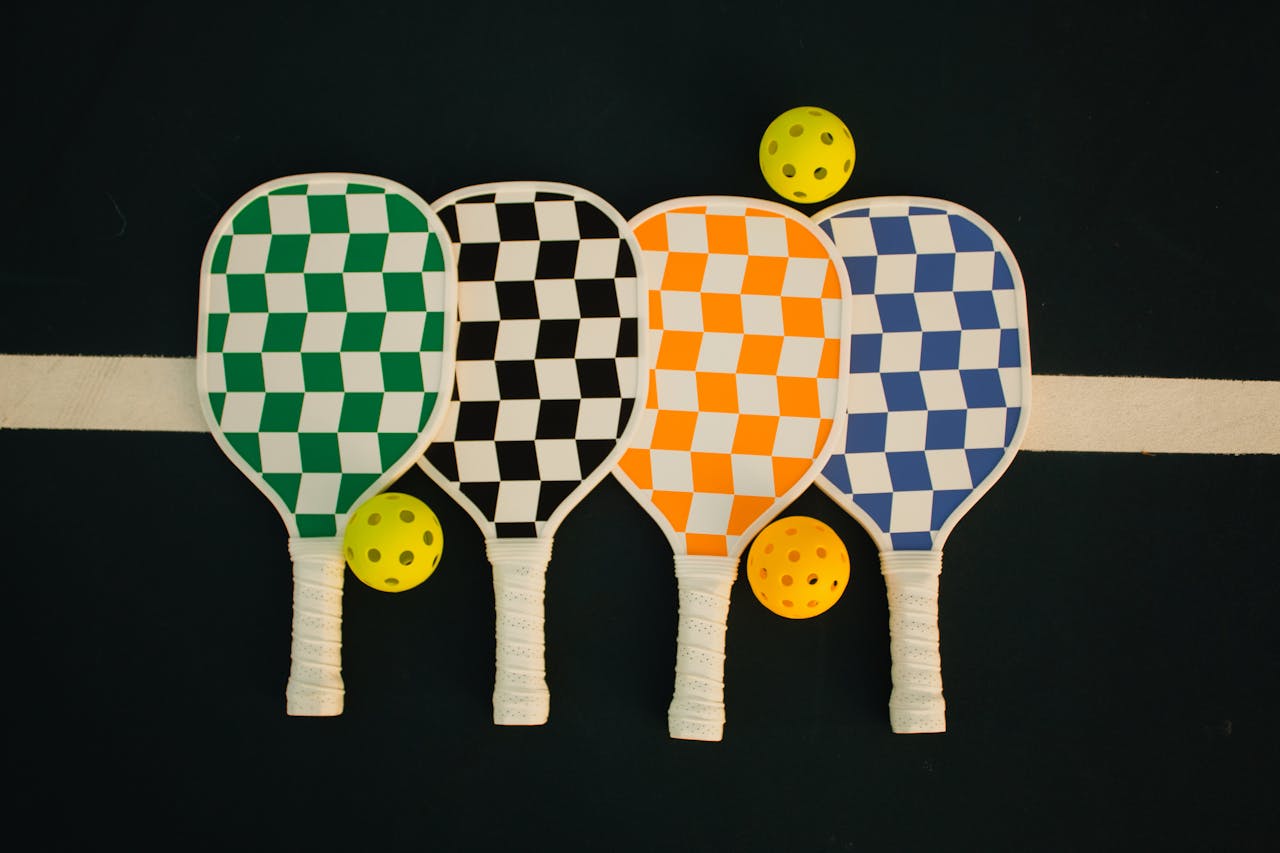 services-01 Four checkerboard-patterned pickleball paddles with wiffle balls on a sports court.