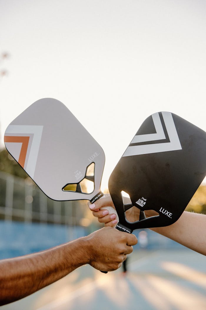 services-03 Two hands holding pickleball paddles outdoors during sunset.